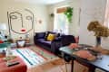 Colorful living room with abstract face wall art, purple sofa, yellow pillow, vibrant rug, and dining table set with pink dishes.