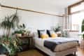 Bright bedroom with a bed, yellow and gray pillows, surrounded by lush green plants and an open window.