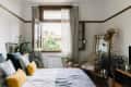 Cozy bedroom with large window, plants, wooden furniture, and a bed with blue and yellow pillows.