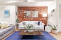 Living room with exposed brick wall, white sofa, blue rug, glass coffee table, and modern decor elements.