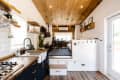 Modern tiny house interior with wood ceiling, black cabinets, open shelves, and a raised living area with a drum set.