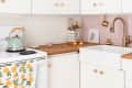 Cozy kitchen with white cabinets, brass faucet, pink backsplash, mint kettle, and citrus-patterned towel on stove.