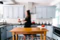 Woman in a kitchen with light blue cabinets, white subway tiles, and a wooden island with tomatoes on a cake stand.