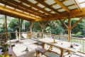 Covered wooden patio with a hammock, dining table, and a cat, surrounded by lush greenery.