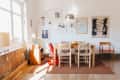 Dining room with wooden table, woven chairs, red high chair, wall art, and potted plants by a window.
