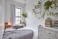 Cozy bedroom with a bed, gray knit blanket, books, potted plants, and hanging greenery on white walls.