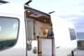 Camper van with open sliding door, showing interior kitchen, plants, and ocean view in the background.