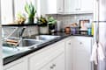 Bright kitchen corner with white cabinets, stainless steel sink, dish rack, potted plants, and colorful bowls on the counter.