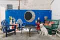 Patio with blue chairs, red tables, a fire pit, mosaic wall art, potted plants, and a white dog sitting on the ground.