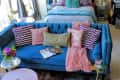 Blue velvet sofa with patterned pillows, pink throw, and open magazine in a cozy living room with a bed in the background.