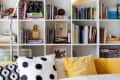 Bookshelf with colorful books, modern lamp, yellow sofa, and patterned cushions in a cozy living room.