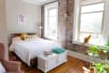 Bedroom with brick wall, bed with "Merci" pillow, cat on windowsill, plants, and books on a white chest.
