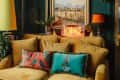 Cozy living room with mustard sofa, colorful cushions, neon "Lounge" sign, and eclectic decor.