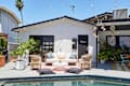 Poolside patio with white sofa, wicker chairs, potted plants, and string lights against a white building.