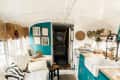 Cozy converted bus interior with teal cabinets, wicker decor, a farmhouse sink, and a cushioned seating area with plants.
