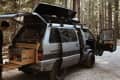 Camper van with open doors and kitchen setup in a forest, person standing nearby.