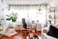 Home office with white desk, black chair, large plant, teepee, and gold floor lamp. Bookshelf and sofa with black pillow.