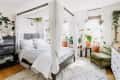 Canopy bed with white curtains, surrounded by potted plants, green velvet chair, and a dresser in a bright bedroom.