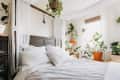 Bedroom with white bedding, wooden headboard, and numerous potted plants by a window with sheer curtains.