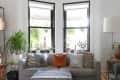 Gray sofa with assorted pillows, surrounded by potted plants, under large windows in a bright living room.