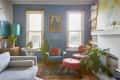 Cozy living room with blue walls, vintage armchair, pink ottomans, plants, bookshelves, and a framed portrait.