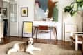 Living room with abstract art, a brown chair, a desk, and a dog on a woven rug. Large plant in the corner.