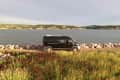 Black camper van parked by a lake with rocky shore, surrounded by grassy fields and distant hills.