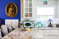 A small square art print above a kitchen window in a white-walled kitchen.