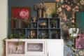 White cabinet with glassware and plates, surrounded by plants, art, and floral wallpaper, next to a red armchair.