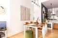 Dining area with wooden table, mustard chairs, geometric wall art, and view into a modern kitchen with plants and open shelving.