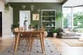 Dining room with wooden table, wicker chairs, green wall, plant shelves, and a cozy sofa with a dog by large windows.