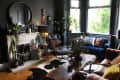 Cozy living room with dark walls, blue velvet sofa, leather chair, plants, and a wooden coffee table.