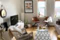 Cozy living room with gray sofa, wicker chair, two brown chairs, TV, plants, and geometric rug under a glass coffee table.