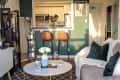 Cozy living room with beige sofa, green pillows, round coffee table, and view into kitchen with bar stools and flowers.