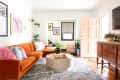 Bright living room with orange sofa, colorful pillows, round coffee table, and eclectic wall art.