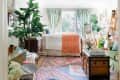 Cozy bedroom with green curtains, patterned rugs, plants, and a wooden chest. Bed with orange throw and art on easel.