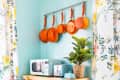 Kitchen corner with hanging orange pans, floral curtains, wooden cart with microwave, toaster, plant, and colorful decor.