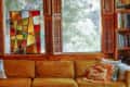 Cozy living room with mustard sofa, colorful pillows, stained glass window, wooden shutters, and bookshelves.