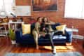 Couple sitting on a blue sofa with a black dog, surrounded by yellow pillows, in a room with brick walls and wooden floors.