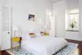 Bright bedroom with white bedding, yellow side tables, lamps, colorful artwork, and a plant on the windowsill.
