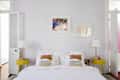 Bedroom with white bedding, colorful pillows, yellow side tables, and two lamps. Art hangs above the bed.