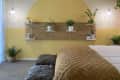 Bedroom with yellow accent wall, floating shelves, potted plants, brown quilted bedspread, and ceiling fan.