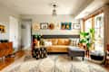 Living room with tan sofa, colorful pillows, wall art, large plants, and a patterned rug near sliding glass doors.