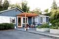 Blue house with orange door, wooden pergola, white chairs, potted plants, and raised garden beds in front yard.