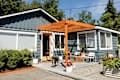 Single-story blue house with a wooden pergola, patio chairs, potted plants, and a dog on the front porch.