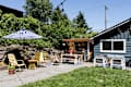 Patio with yellow chairs, striped umbrella, fire pit, and outdoor dining area next to a blue house with string lights.
