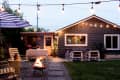 Cozy backyard patio with string lights, fire pit, striped umbrella, and outdoor seating in front of a gray house.