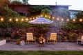 Patio with two yellow chairs, a striped umbrella, string lights, and a fire pit against a stone wall with ivy.