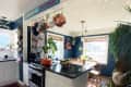 Kitchen with dark blue walls, hanging pots, plants, and a dining nook with wooden chairs and a window.