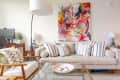 Beige sofa with striped cushions, abstract colorful painting, glass coffee table, and modern floor lamp in a living room.